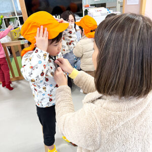 保育士に防災用の帽子を被せてもらう子ども