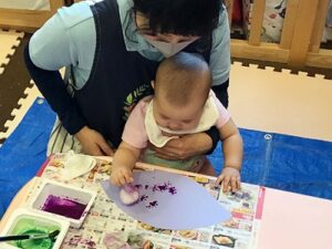 保育士に抱きかかえられながら絵の具のついたお手製スタンプを押す子ども